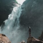 man standing on brown rock cliff in front of waterfalls photography